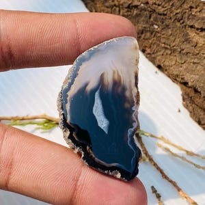 May include: A polished agate stone slice with a natural, irregular shape. The stone features bands of black, white, and brown hues, with a central white inclusion. The edges have a rough, textured appearance.