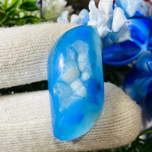 Könnte beinhalten: Ein polierter, oval geformter Edelstein in leuchtendem Blau. Der Stein weist einen zentralen Bereich mit weißen, kristallinen Formationen auf, was einen auffälligen Kontrast erzeugt. Der Edelstein wird von einer behandschuhten Hand gehalten, mit floralen Akzenten im Hintergrund.