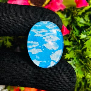May include: An oval cabochon featuring a sky-blue design with white cloud patterns. The gemstone is set against a backdrop of pink flowers and green foliage. A unique piece of jewellery.