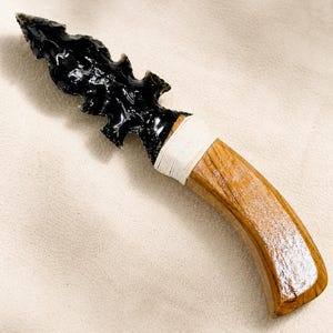 May include: A knife featuring a serrated black obsidian blade attached to a curved, polished wooden handle. The connection point between the stone blade and the wood is secured with tightly wound white cord, creating a distinct contrast in texture.