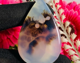 May include: A teardrop-shaped gemstone with a landscape pattern in white, blue, brown, and purple. The polished stone has a translucent quality and a smooth surface. The gemstone is set against a backdrop of pink flowers and a black surface.