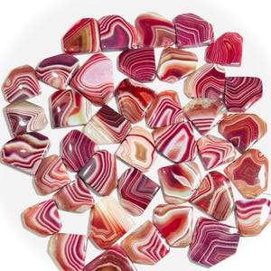 May include: A collection of polished, irregularly shaped agate stones featuring vibrant bands of red, pink, white, and deep burgundy. Each stone displays unique concentric layers and smooth, glossy surfaces, arranged in a loose, scattered cluster.