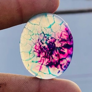 May include: Oval gemstone with a distinctive pattern of white, teal, pink, and purple hues. The stone features a cracked, web-like design. The gemstone is held between two fingers.