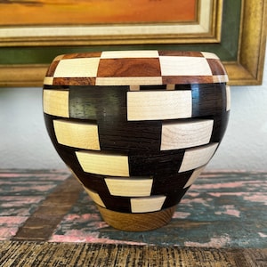 May include: A wooden bowl with a checkered pattern in shades of brown, black, and white. The bowl is made of different types of wood, creating a unique and intricate design.