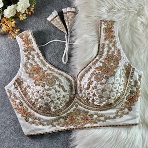 May include: A sleeveless white blouse featuring intricate gold embroidery, sequins, and floral patterns in shades of orange and green. The garment has a deep V-neckline, a structured bodice with a grid-like sequin design, and white tie-up back straps.