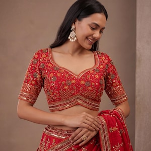 May include: A vibrant red blouse featuring intricate gold embroidery, floral patterns, and dense sequin embellishments. The garment has a sweetheart neckline, short sleeves, and a structured hemline, paired with a matching red skirt with embroidery.