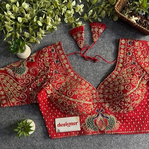 May include: A red blouse featuring intricate gold embroidery, sequin detailing, and peacock motifs. The garment has a deep V-neckline, short sleeves, and a back tie closure with two triangular tassels. A white tag with the word designer is attached.