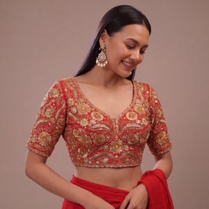 May include: A red blouse featuring a deep V-neckline and short sleeves, adorned with intricate gold floral embroidery, metallic thread work, and small sequins throughout the fabric. The garment has a fitted silhouette and a scalloped hemline detail.