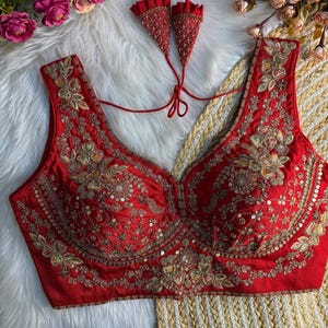 Peut inclure: Un haut sans manches rouge avec des broderies dorées complexes et des paillettes. Le haut présente un décolleté en V profond et est orné de motifs floraux et cachemire. Deux pompons rouges perlés assortis sont attachés.