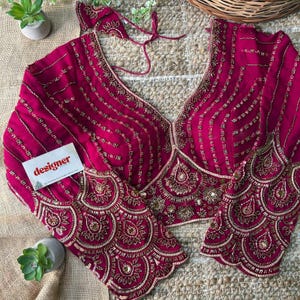 Puede incluir: Una blusa de color magenta con intrincados abalorios dorados. La blusa presenta un escote en V profundo y mangas largas con puños acampanados. El diseño incluye patrones verticales y circulares de cuentas doradas. Se ve una pequeña tarjeta con la palabra "designer".