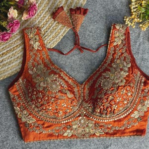 Puede incluir: Una blusa sin mangas de color naranja óxido con un escote en V profundo y un intrincado bordado dorado con lentejuelas. La prenda incluye un cierre de lazo en la espalda con dos borlas de tela triangulares adornadas con bordado dorado.