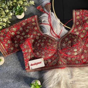 May include: A red, intricately embroidered top with gold and copper-colored embellishments. The top features a deep V-neck and short sleeves. A small card with the word "designer" is visible.