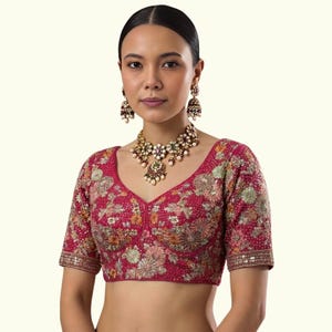 Op de afbeelding: Een magenta geborduurde blouse met een V-hals en korte mouwen. De blouse heeft ingewikkelde bloempatronen in goud, groen en oranje. Een bijpassende ketting en oorbellen maken de set compleet, met een traditioneel ontwerp.