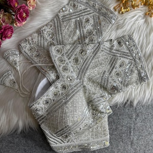 May include: A white, heavily embellished top with silver sequins and beads. The intricate design features geometric patterns and floral accents. The top is likely for special occasions, with a luxurious and ornate appearance.