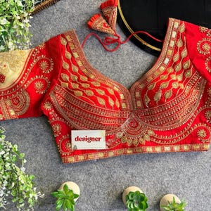 Puede incluir: Una blusa roja con intrincados bordados dorados. La blusa presenta un escote en V profundo y detalles ornamentados a lo largo del escote, las mangas y el corpiño. La palabra "designer" está impresa en una pequeña tarjeta blanca.