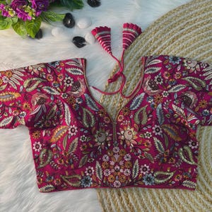 Puede incluir: Una blusa magenta de manga corta con intrincados bordados florales. La blusa presenta un escote en V profundo y borlas decorativas. El bordado incluye varios colores, como dorado, verde y azul, creando un diseño detallado y ornamentado.