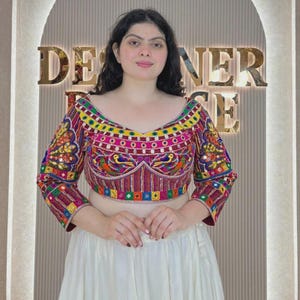 May include: A cropped blouse featuring intricate multi-colored embroidery with bird motifs, small mirrors, and geometric patterns. The garment has three-quarter length sleeves, a wide neckline, and a vibrant palette of red, yellow, blue, and green.