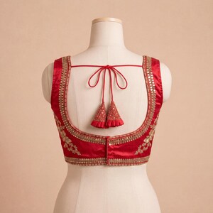 Op de afbeelding: Een rode mouwloze blouse met gouden borduursels en versieringen. De blouse heeft een ronde rug en een striksluiting met decoratieve kwastjes. De halslijn en armsgaten zijn afgewerkt met gouden details. De blouse wordt op een mannequin gepresenteerd.