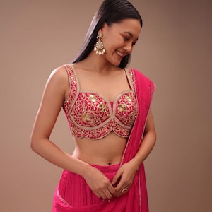 May include: A vibrant pink embroidered top with gold accents and mirror embellishments. The top features a sweetheart neckline and is paired with a matching pink draped garment. The image showcases a traditional Indian garment.