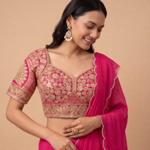 Könnte beinhalten: Eine leuchtend pinkfarbene Bluse mit aufwendiger goldener Stickerei. Die Bluse hat einen quadratischen Ausschnitt und kurze Ärmel. Das Design umfasst florale und geometrische Muster. Ein passender pinkfarbener Schal ist über die Schulter drapiert.