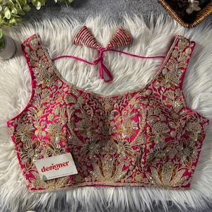 May include: A sleeveless, cerise blouse with detailed gold embroidery and sequin detailing. The blouse has a square neckline and a matching bow. A small tag with the word "designer" is attached.