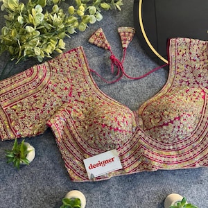 Könnte beinhalten: Ein magenta-farbenes, stark besticktes Oberteil mit goldenen Blumenmustern. Das Kleidungsstück hat einen tiefen Ausschnitt und kurze Ärmel. Das Wort "designer" ist auf einer kleinen weißen Karte aufgedruckt.