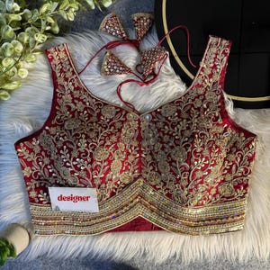 Puede incluir: Una blusa sin mangas, de color rojo intenso, con intrincados bordados dorados y detalles de lentejuelas. La blusa presenta un cuello en V y un borde decorativo. La palabra "designer" está impresa en una pequeña tarjeta blanca. Se incluyen elementos decorativos adicionales a juego.