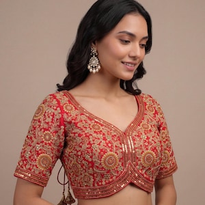 May include: A vibrant red blouse featuring intricate gold embroidery, floral patterns, and sequin embellishments. The garment has a V-neckline, short sleeves, and a scalloped hemline. It is paired with dangling gold earrings with white pearls.