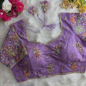May include: A lavender-colored, embroidered blouse with a square neckline. The blouse features intricate floral embroidery in various colors, including gold, red, and green. The sleeves and neckline are adorned with gold trim. Two matching tassels are attached.
