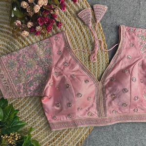 Puede incluir: Una blusa rosa claro con intrincados bordados y adornos. La blusa presenta un escote en V profundo y mangas cortas con patrones florales detallados. Delicados acentos plateados y un ribete decorativo realzan el diseño. La blusa está sobre un fondo neutro.