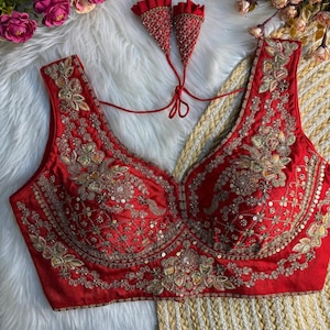 May include: A sleeveless red blouse featuring intricate gold embroidery, sequins, and beadwork throughout the front. The garment has a deep scoop neckline and includes two matching red fabric tassels with gold detailing attached by thin red cords.