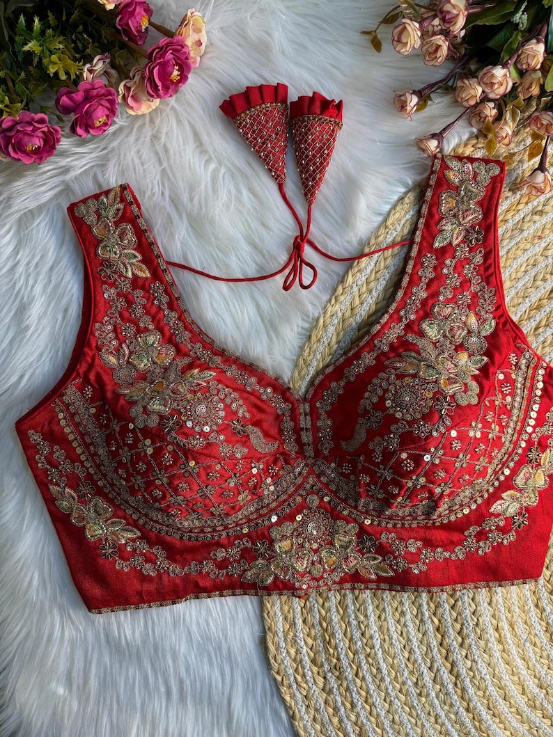 May include: A sleeveless red blouse featuring intricate gold embroidery, sequins, and beadwork throughout the front. The garment has a deep scoop neckline and includes two matching red fabric tassels with gold detailing attached by thin red cords.