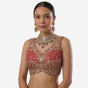 May include: A sleeveless red blouse featuring intricate gold embroidery, sequins, and a V-neckline. The garment is paired with a matching gold-toned statement necklace and dangling jhumka earrings, both adorned with white pearls and red stones.
