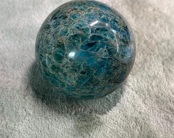 May include: A polished, spherical gemstone with a mottled appearance. The sphere is primarily teal and turquoise, with a network of lighter, crack-like patterns across its surface. The gemstone is smooth and reflective, suggesting a high-quality finish.