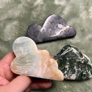 May include: Three cloud-shaped stones in different colours. One is light blue and tan, another is dark grey, and the third is green and white. The stones are smooth and polished, with natural variations in colour and texture.