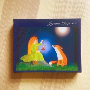 Puede incluir: Caja de rompecabezas con una ilustración caprichosa. La imagen representa a una niña con un vestido verde sosteniendo una linterna, con un zorro mirándola bajo la luna llena. La caja es azul oscuro y tiene el texto "Jigsaw 100 pieces".