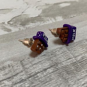 May include: A pair of novelty stud earrings. Each earring is shaped like a miniature chocolate bar with a purple wrapper and the word "MILK" on the side. The chocolate is brown, and the wrapper is purple.