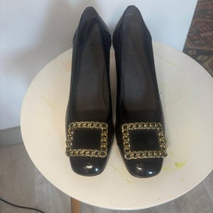 May include: A pair of black pumps featuring a perforated texture on the upper and a square toe. Each shoe is adorned with a prominent, chunky gold-tone metal chain link buckle across the front. The heels are closed and have a smooth finish.
