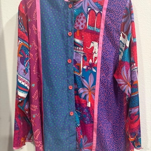 May include: A patterned button-up shirt in a patchwork design. The shirt features a mix of floral, geometric, and paisley patterns in pink, purple, blue, and red. Pink ribbon accents the seams. The shirt has a mandarin collar.