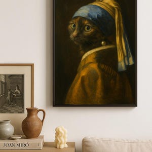 Op de afbeelding: Een ingelijste print van een kat met een gele en blauwe hoofddoek en een parel oorbellen, geïnspireerd op Vermeers "Meisje met de parel". De kat heeft bruin en zwart haar tegen een donkere achtergrond. De print hangt boven een houten bijzettafel.