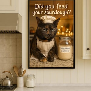 May include: A framed print in a kitchen setting features a cat in a chef's hat and apron, with the text "Did you feed your sourdough?" and a jar labeled "Sourdough Starter". The kitchen has cream-colored cabinets and a marble countertop.