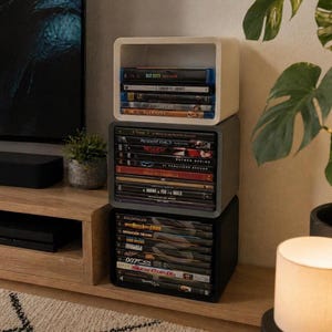 Op de afbeelding: Drie gestapelde vierkante opbergunits in wit, grijs en zwart, gevuld met dvd's en Blu-ray discs. De bovenste unit is wit, de middelste is grijs en de onderste is zwart. De schijven hebben kleurrijke covers en titels zoals "Bad Boys".