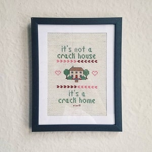 May include: A framed cross-stitch with the text "It's not a crack house, it's a crack home." The cross-stitch features a small house with green trees and a red door.