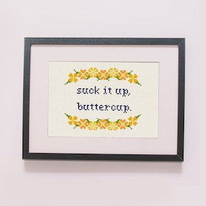 May include: A framed cross-stitch with a floral border and the text "suck it up, buttercup."