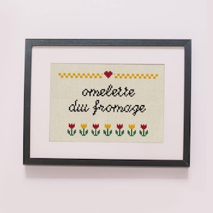 Puede incluir: Obra de arte en punto de cruz enmarcada con la frase "omelette du fromage" en escritura negra. El diseño incluye un borde amarillo, un corazón rojo y una fila de tulipanes rojos, amarillos y verdes. El marco es negro.