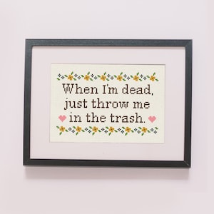 May include: A framed cross-stitch with a white background and brown text that reads "When I'm dead, just throw me in the trash." The text is surrounded by a border of yellow and blue flowers. Two pink hearts are on either side of the text.