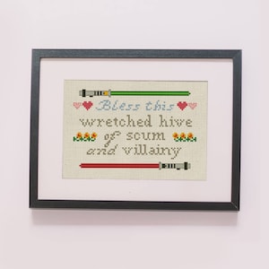 May include: A framed cross-stitch design with a Star Wars theme. The design features two lightsabers, hearts, and the text "Bless this wretched hive of scum and villainy."