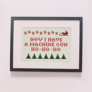 May include: A framed cross-stitch design with a white background and red and green text. The text reads "NOW I HAVE A MACHINE GUN HO-HO-HO". There are green trees and a red sleigh with Santa Claus and reindeer at the top of the design.
