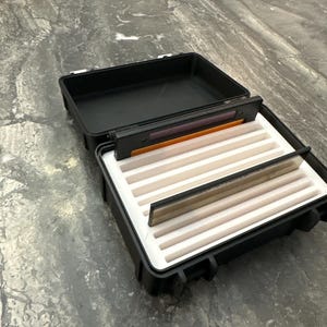 Pu&ograve; includere: Una scatola da pesca a mosca nera aperta con interno in schiuma bianca e scomparti multipli. La scatola contiene varie mosche da pesca a mosca ed &egrave; progettata per proteggere e organizzare le mosche. La scatola &egrave; su una superficie grigia.