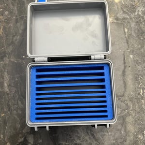 Puede incluir: Una caja de almacenamiento rectangular gris abierta con un interior azul. La caja tiene un inserto de espuma azul con múltiples ranuras horizontales. La caja tiene cierres y bisagras azules. La caja está diseñada para proteger y organizar objetos pequeños.